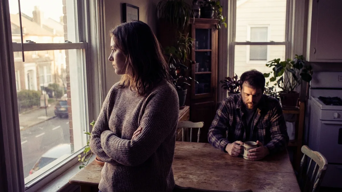 A woman standing at a bright kitchen window with arms crossed, looking out, while a man sits at the table behind her with both hands around a mug, both quietly in the same room