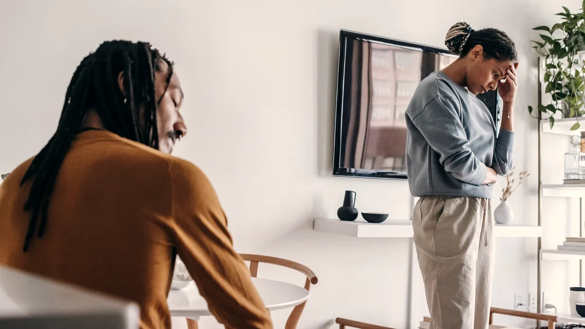 A couple standing apart in a living room after an argument, one seated looking down, the other standing with head bowed, natural diffuse light
