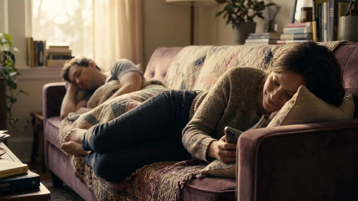 Woman on a mauve sofa in morning light, looking at her phone, partner asleep behind her
