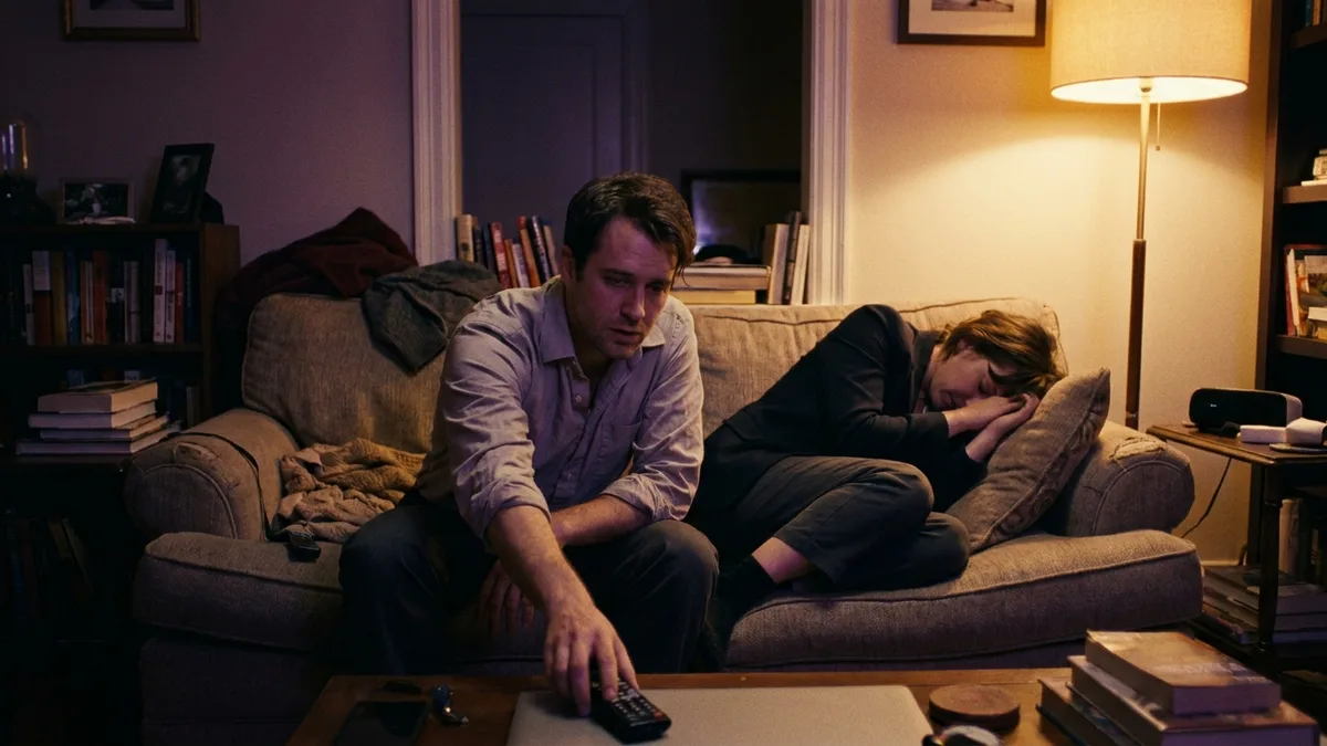 A couple slumped on the sofa late at night in amber lamplight, one reaching for the remote while the other rests with eyes closed